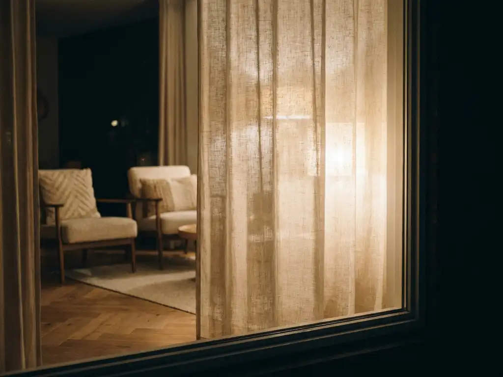 Linnen privacygordijn filtert warm amberkleurig lamplicht in Nederlandse woonkamer met bouclékussens en eiken visgraatparket.