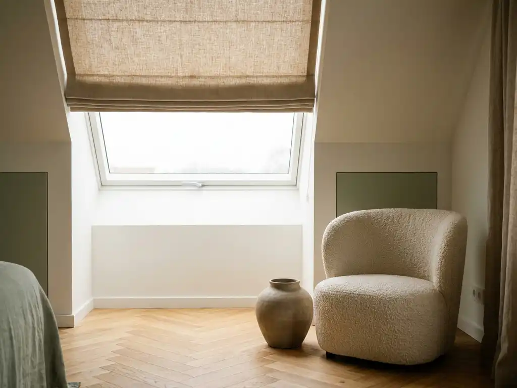 Linnen rolgordijn halfopen voor groot dakraam, zacht noorderlicht op visgraateikenhouten vloer, bouclé fauteuil en matte keramieken vaas op de voorgrond.