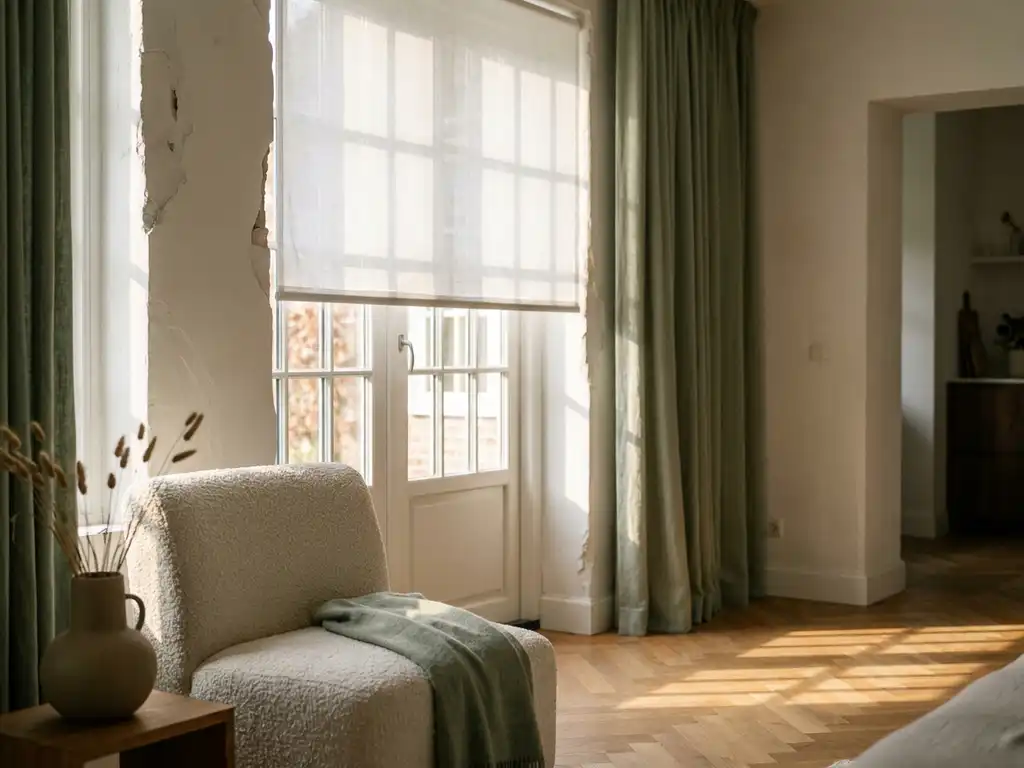 Witte linnen rolgordijn halfopen voor hoog Hollands raam, middaglicht filtert door stof op visgraateikenhouten vloer, bouclé fauteuil en keramische vaas op achtergrond.